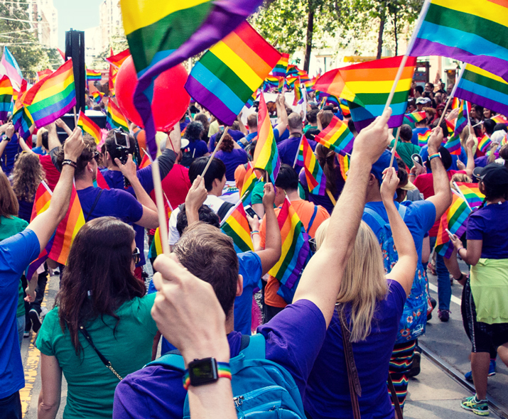 Foto de uma multidão de pessoas em uma parada agitando bandeiras de arco-íris. 