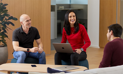 Śmiejąca się Camila siedząca z MacBookiem na kolanach między dwójką innych pracowników Apple.