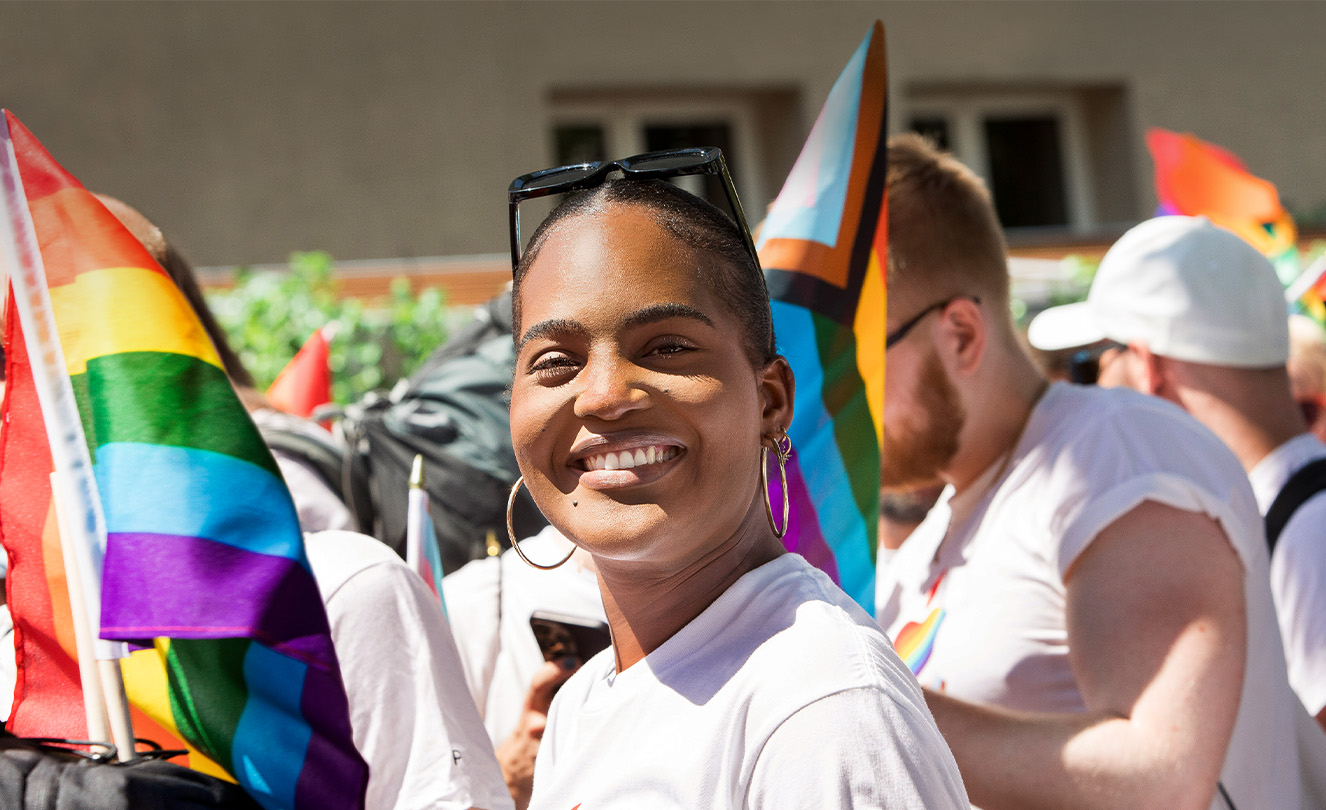 Uśmiechnięta pracownica Apple podczas wydarzenia Apple Pride w tłumie pracowników trzymających tęczowe flagi i ubranych w koszulki z flagą. 